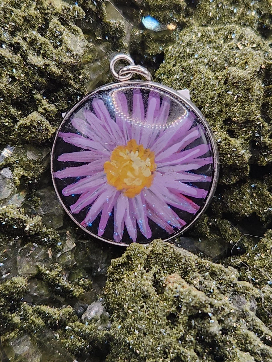 Purple daisy flower art pendant displayed among green crystals. Crystal display and styling props not included.