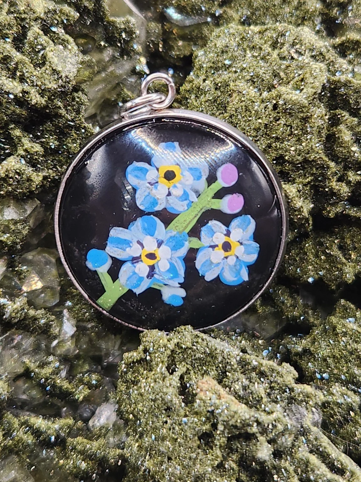 Blue forget-me-not flower art pendant nestled in a sparkling green crystal display. Crystal and display props not included.
