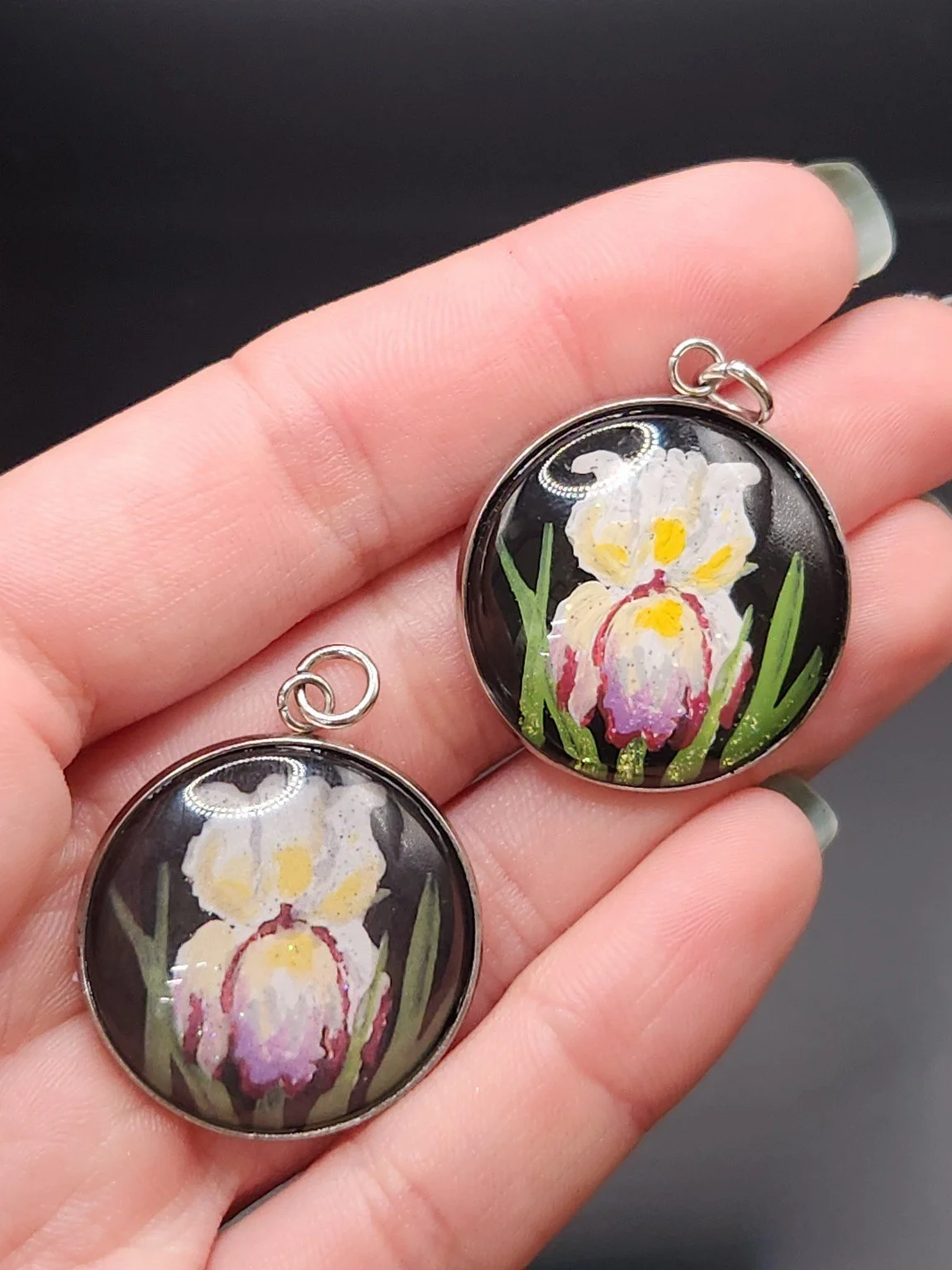 Two iris flower pendants held in a hand: hand-painted original at the top right and printed reproduction at the bottom left.