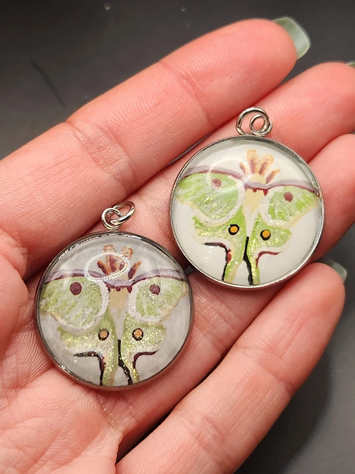 Luna moth pendant comparison held in a hand: the hand-painted original is at the top right, and the printed reproduction is at the bottom left.