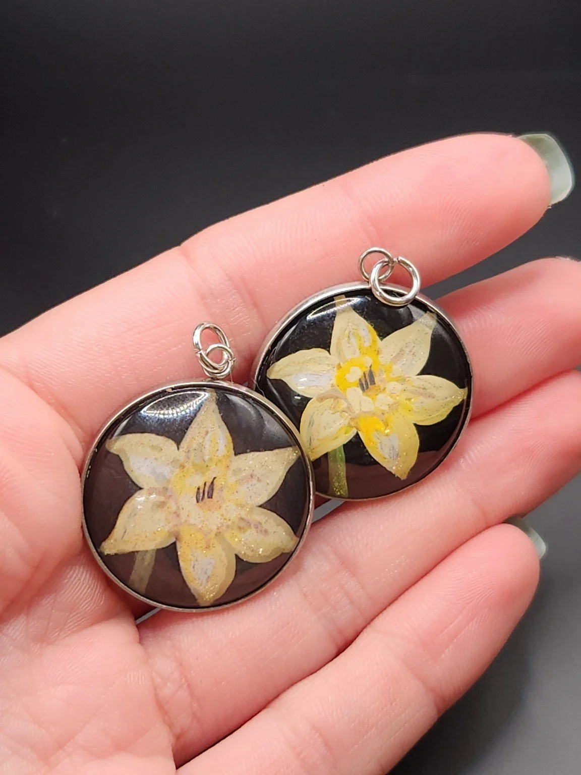 Two daffodil flower pendants held in a hand: hand-painted original at the top right and printed reproduction at the bottom left.