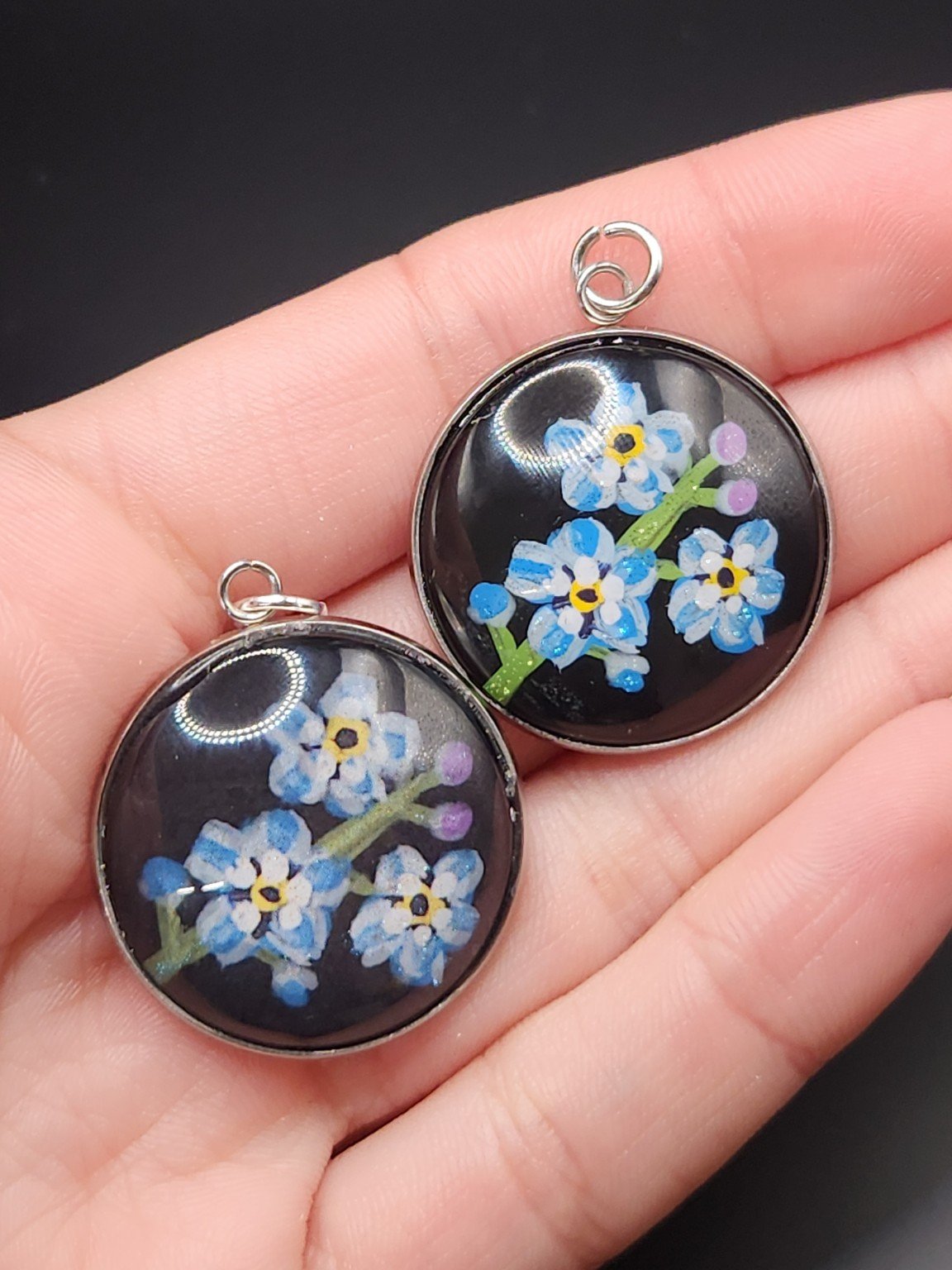 Two blue forget-me-not pendants held in a hand: hand-painted original at the top right and printed reproduction at the bottom left.