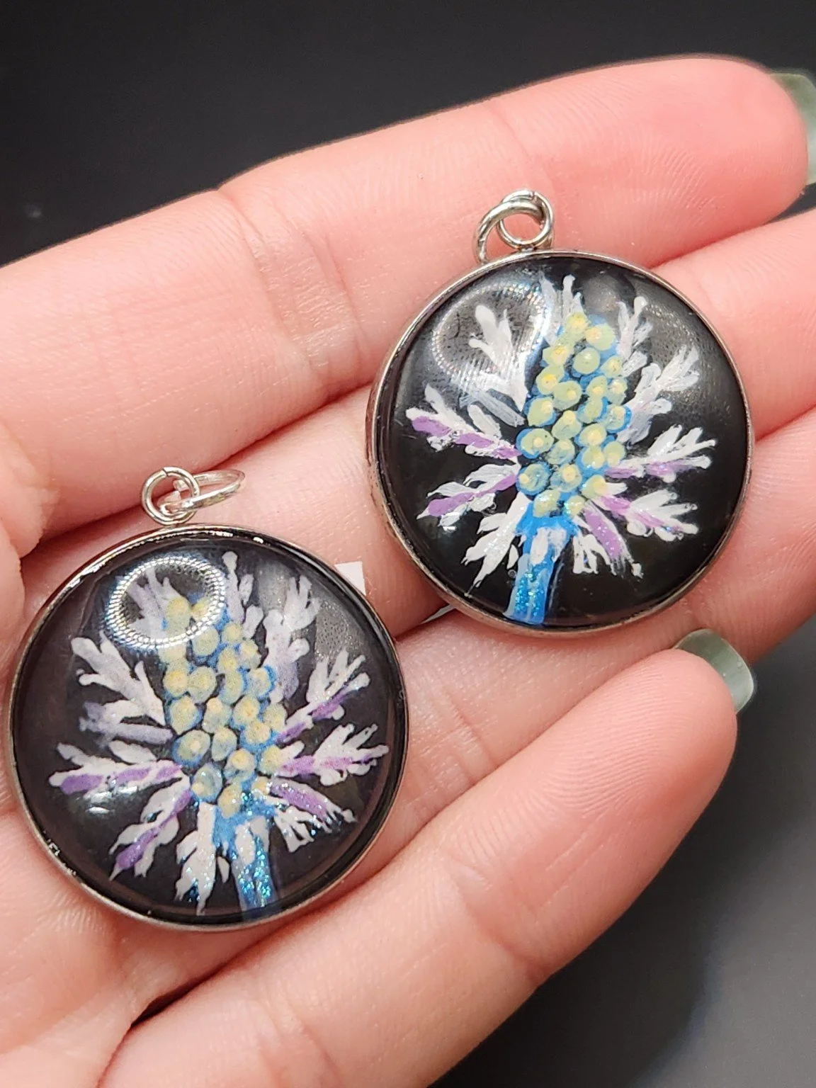 Two eryngium flower pendants in a hand: hand-painted original at the top right and printed reproduction at the bottom left.