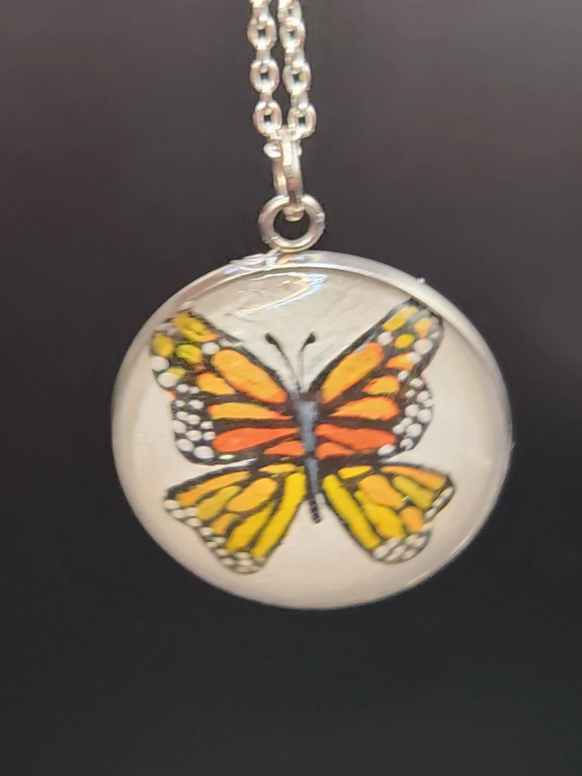Hand-painted orange and black monarch butterfly art pendant hanging from a silver-tone chain against a dark background. Chain not included.