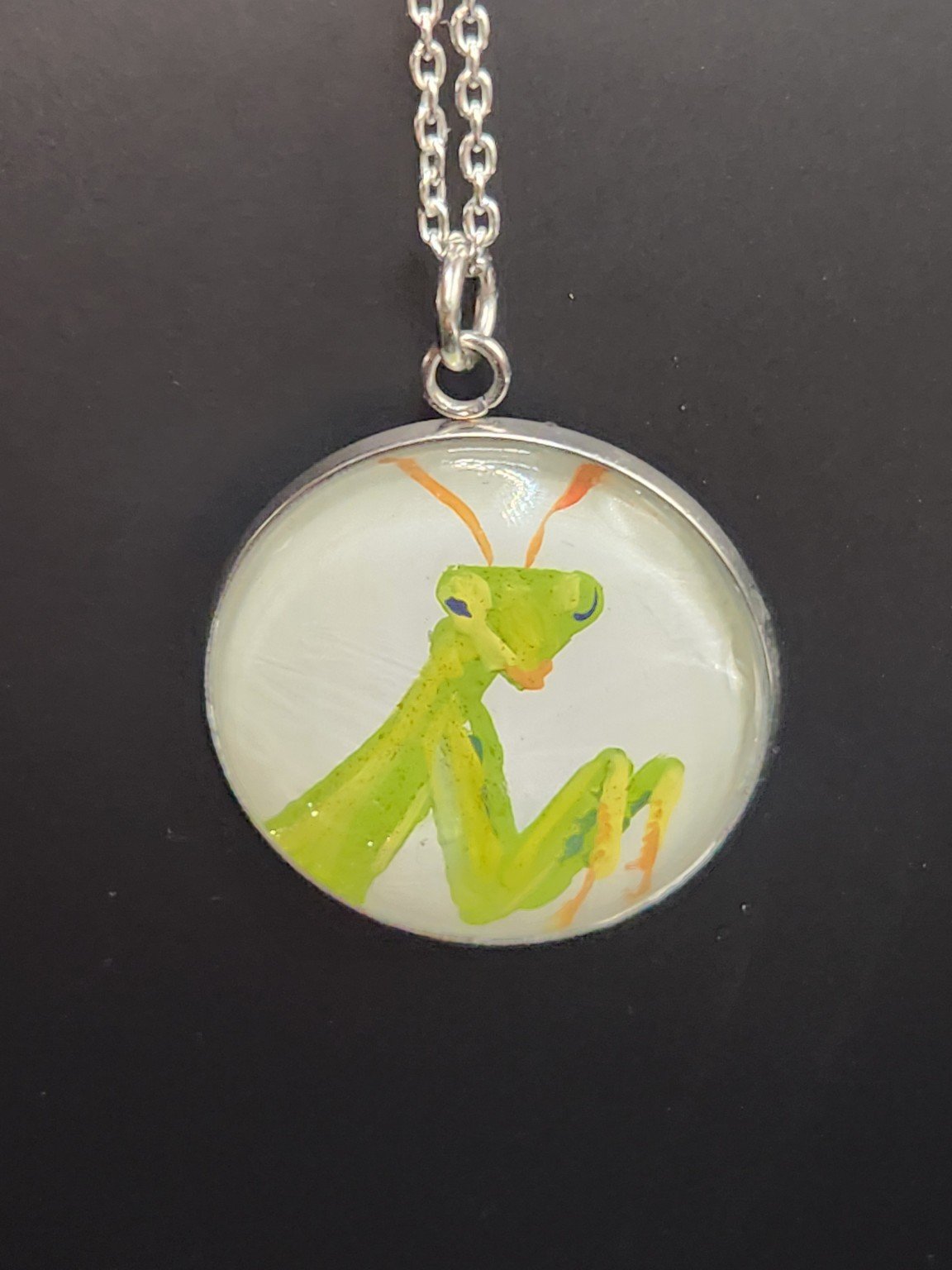 Green praying mantis art pendant shown on a silver-tone chain against a dark background. Chain not included.