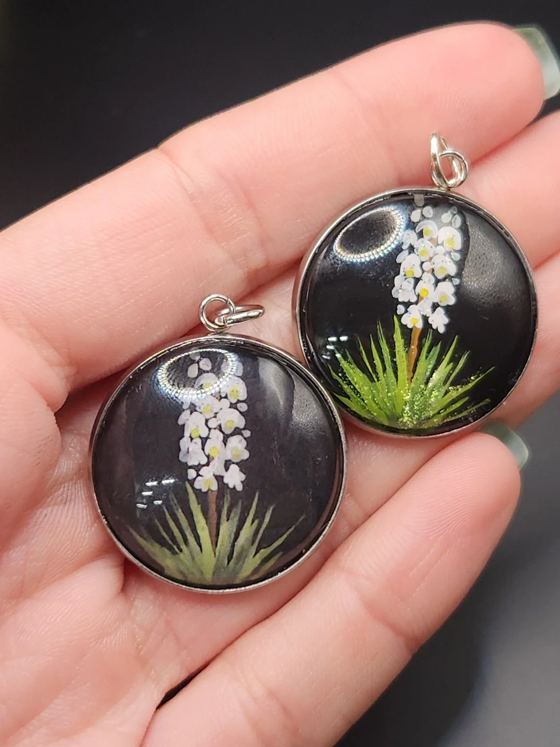 Two yucca flower pendants held in a hand: hand-painted original at the top right and printed reproduction at the bottom left.
