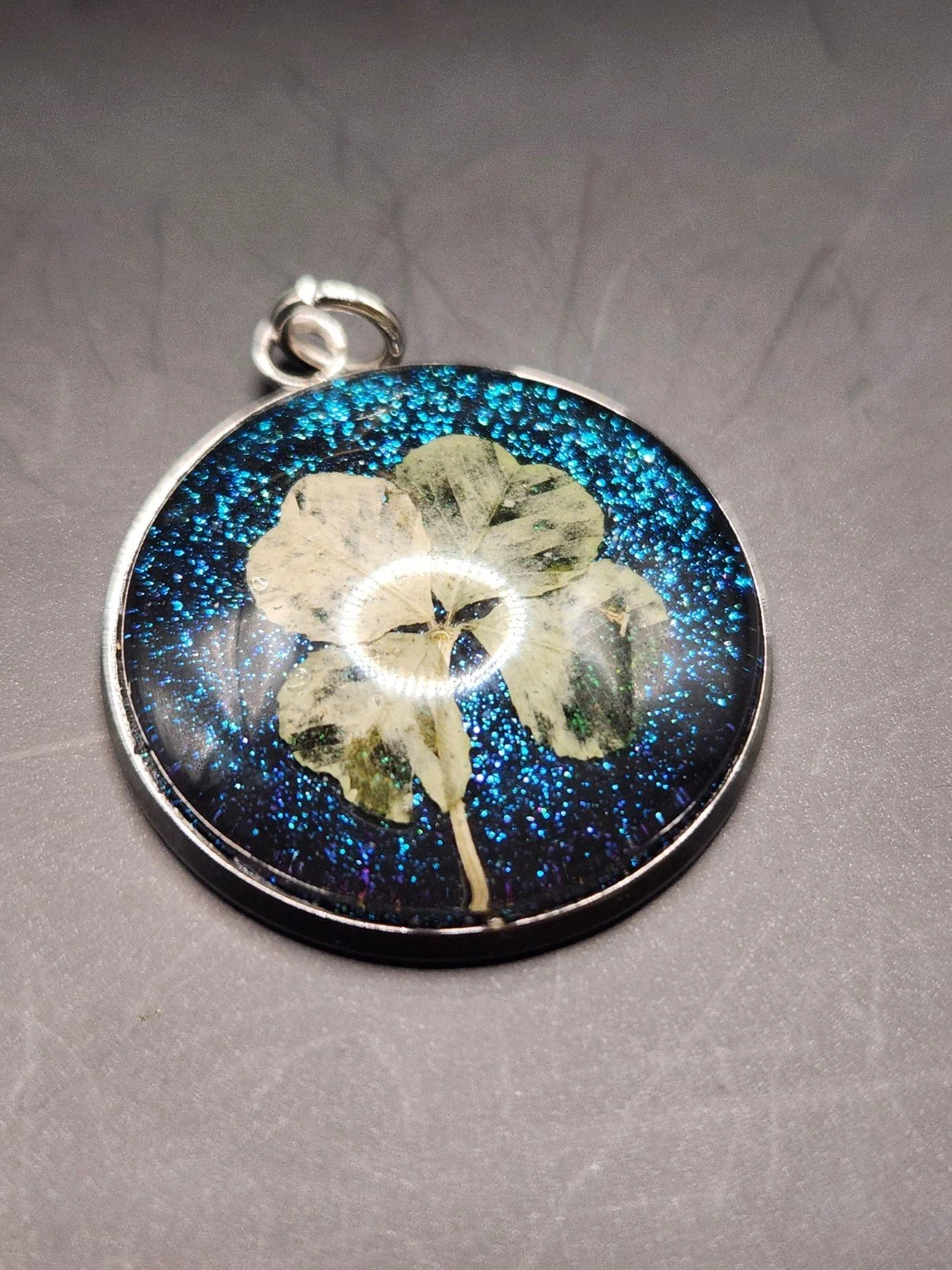 Naturally Unique N004: real pressed four-leaf clover pendant with blue glitter background, close-up on gray.