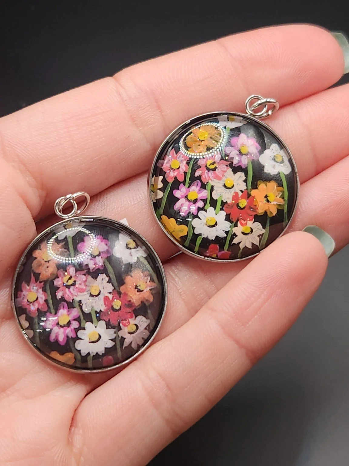 Two cosmos flower pendants held in a hand: hand-painted original at the top right and printed reproduction at the bottom left.
