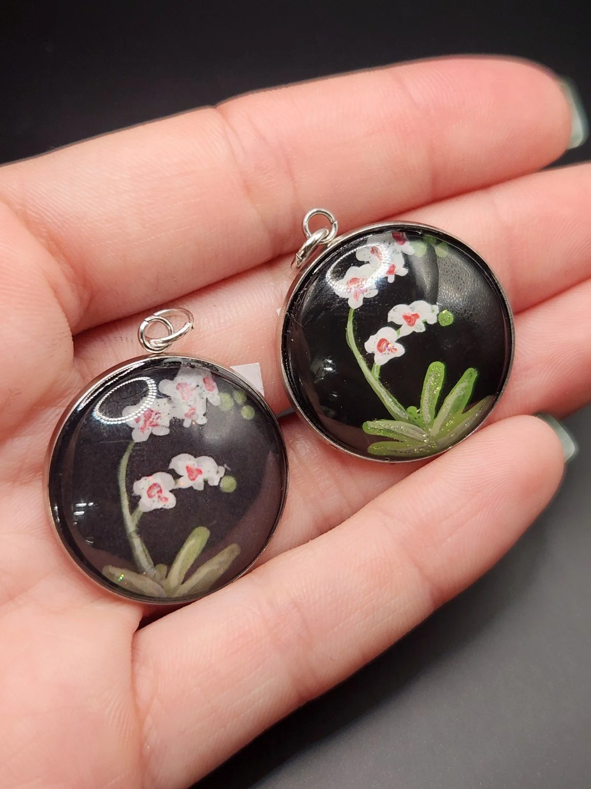 Two orchid flower pendants held in a hand: hand-painted original at the top right and printed reproduction at the bottom left.