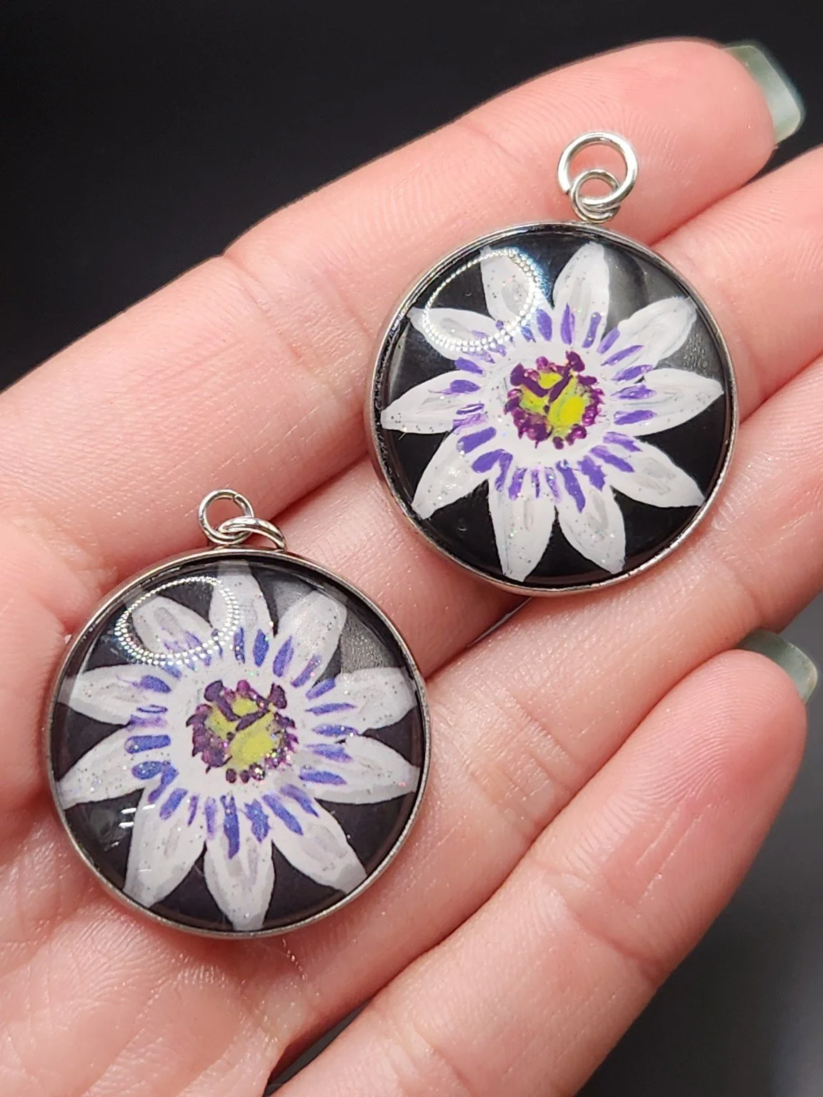 Two passion flower pendants held in a hand: hand-painted original at the top right and printed reproduction at the bottom left.