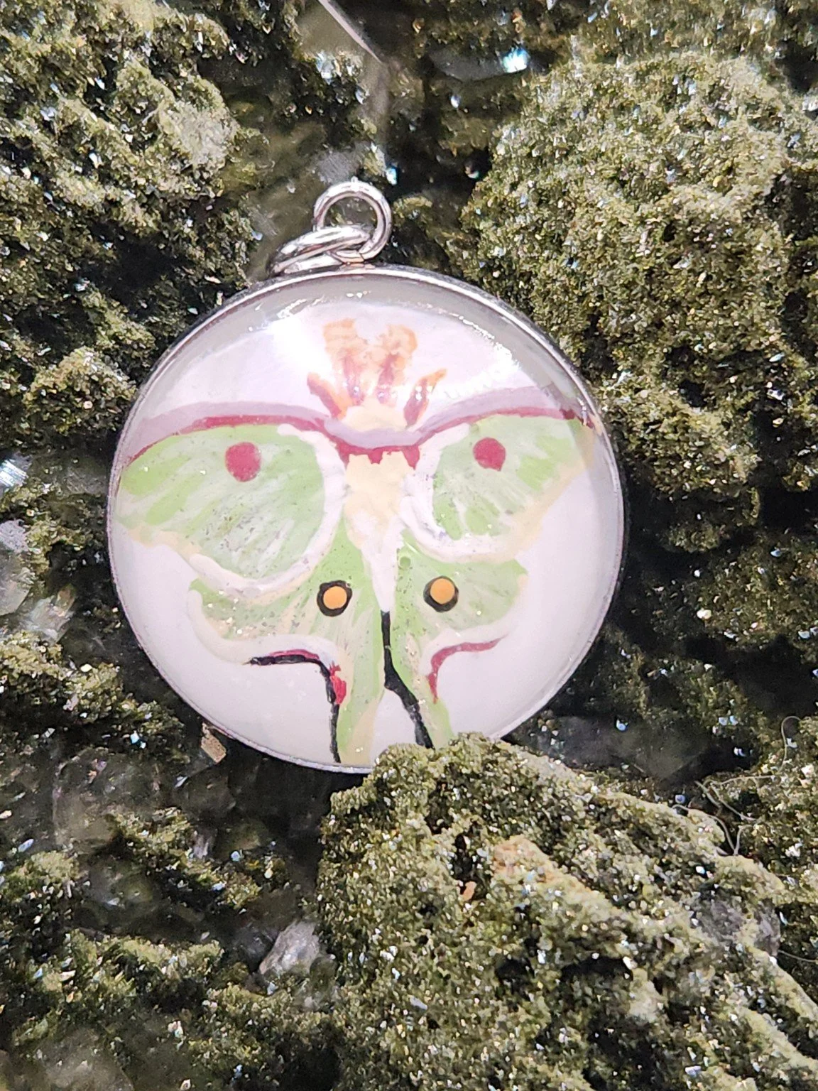 Pale green luna moth art pendant resting on a green crystal display. Crystal display not included.