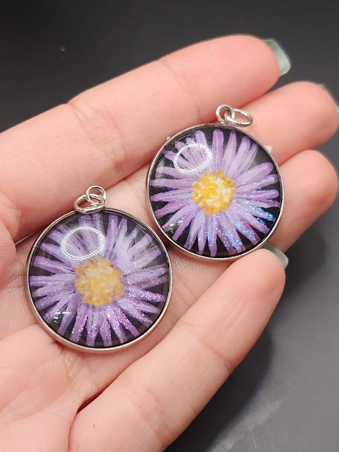 Two purple daisy pendants held in a hand: hand-painted original at the top right and printed reproduction at the bottom left.