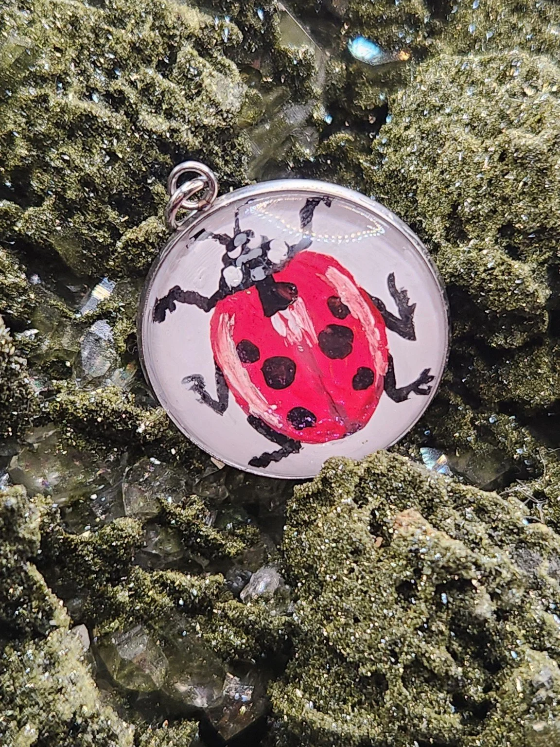 Red and black ladybug art pendant resting on a green crystal display. Crystal display not included.