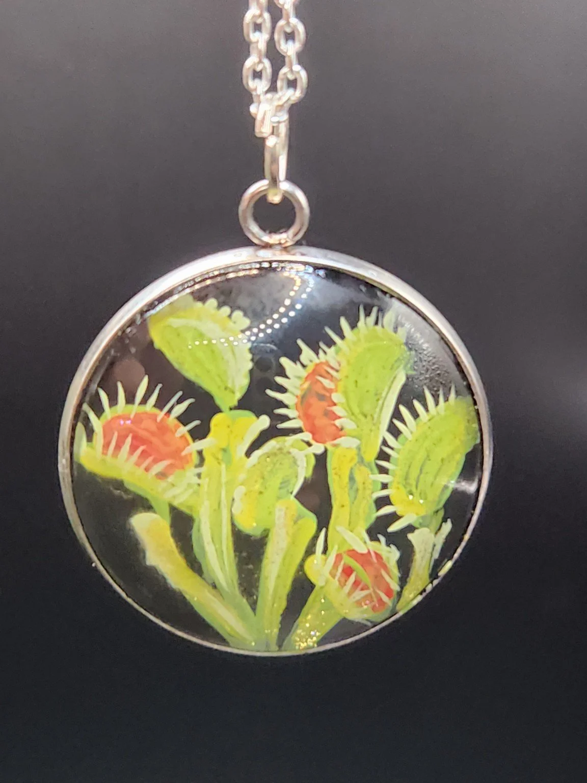 Green and red Venus flytrap art pendant hanging from a silver-tone chain against a dark background. Chain not included.