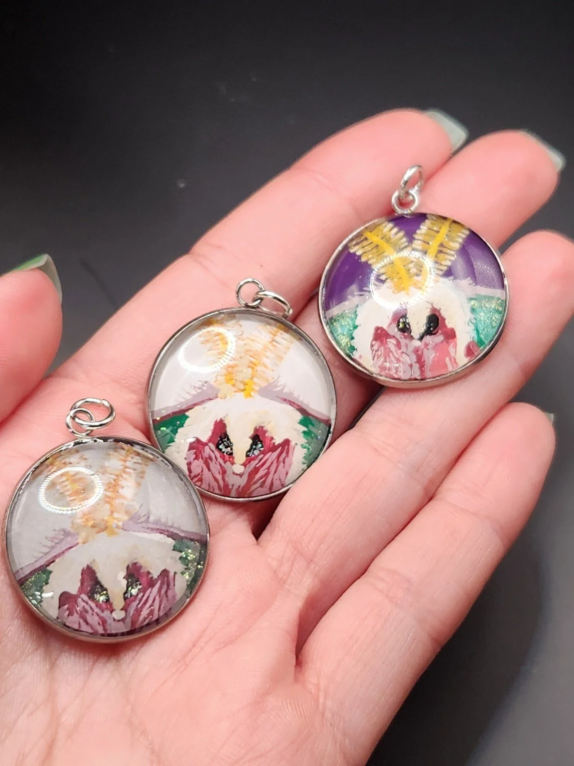 Three luna moth face pendants held in a hand: printed reproduction at far left, white-background hand-painted original in the middle, and purple-background hand-painted original at far right.