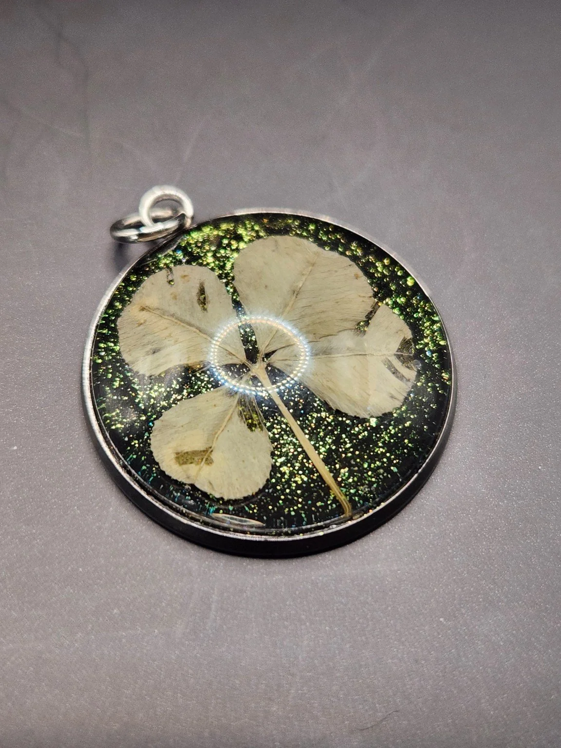Pristine P001: real pressed four-leaf clover pendant with green glitter background, close-up on gray.