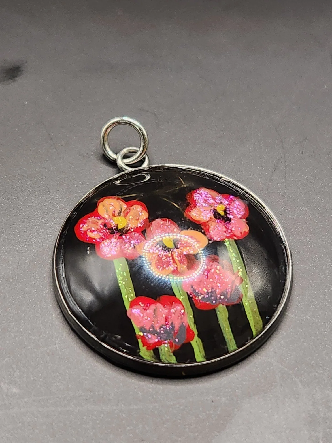 Red and pink poppy flower art pendant with green stems and shimmering glitter in a silver-tone bezel against a gray background.