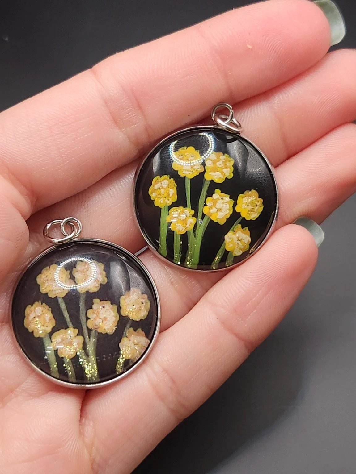 Two billy button flower pendants held in a hand: hand-painted original at the top right and printed reproduction at the bottom left.