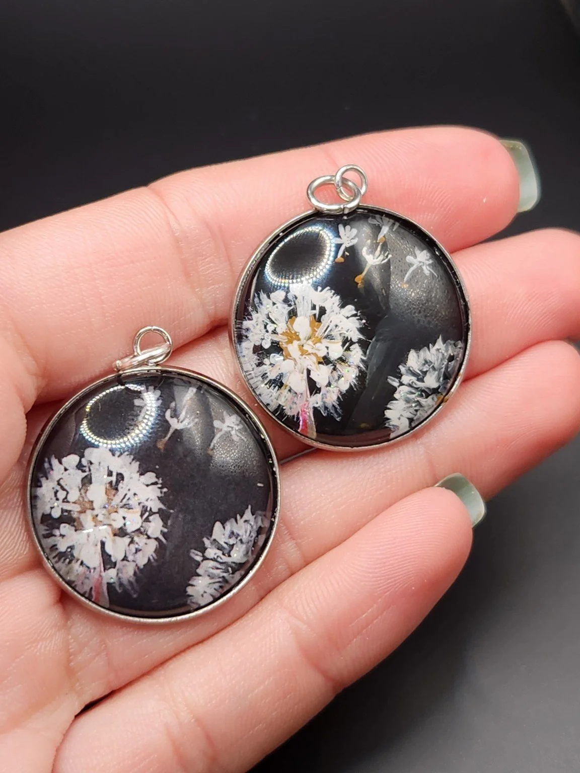 Two Dandelion Wish pendants held in a hand: hand-painted original at the top right and printed reproduction at the bottom left.