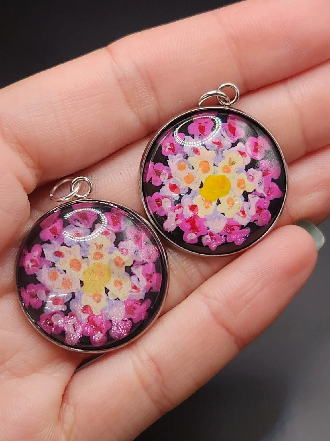 Two lantana flower pendants held in a hand: hand-painted original at the top right and printed reproduction at the bottom left.