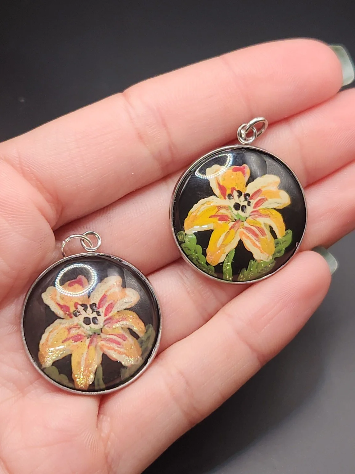 Two daylily flower pendants held in a hand: hand-painted original at the top right and printed reproduction at the bottom left.