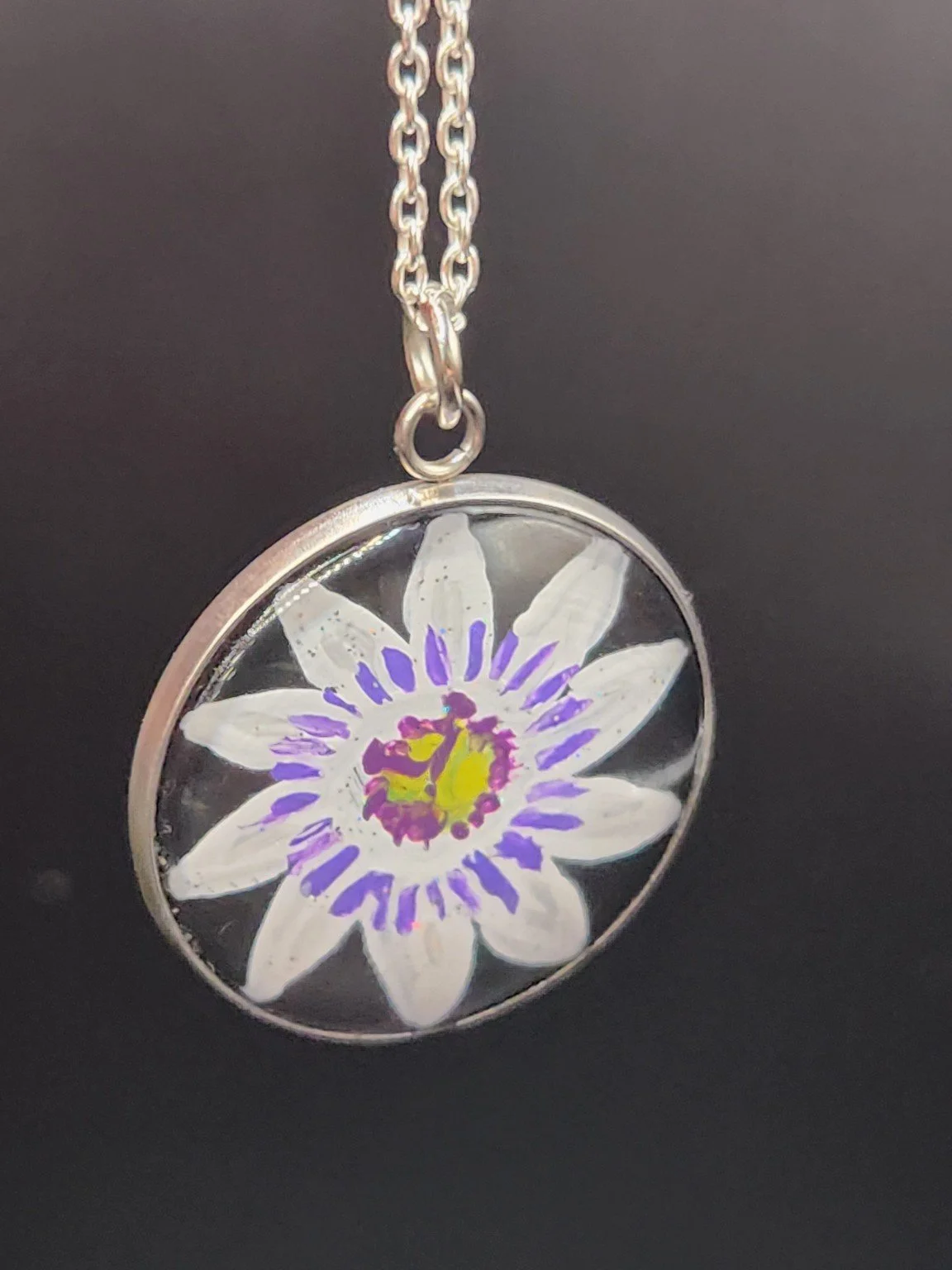 Purple and white passion flower art pendant hanging from a silver-tone chain against a dark background. Chain not included.