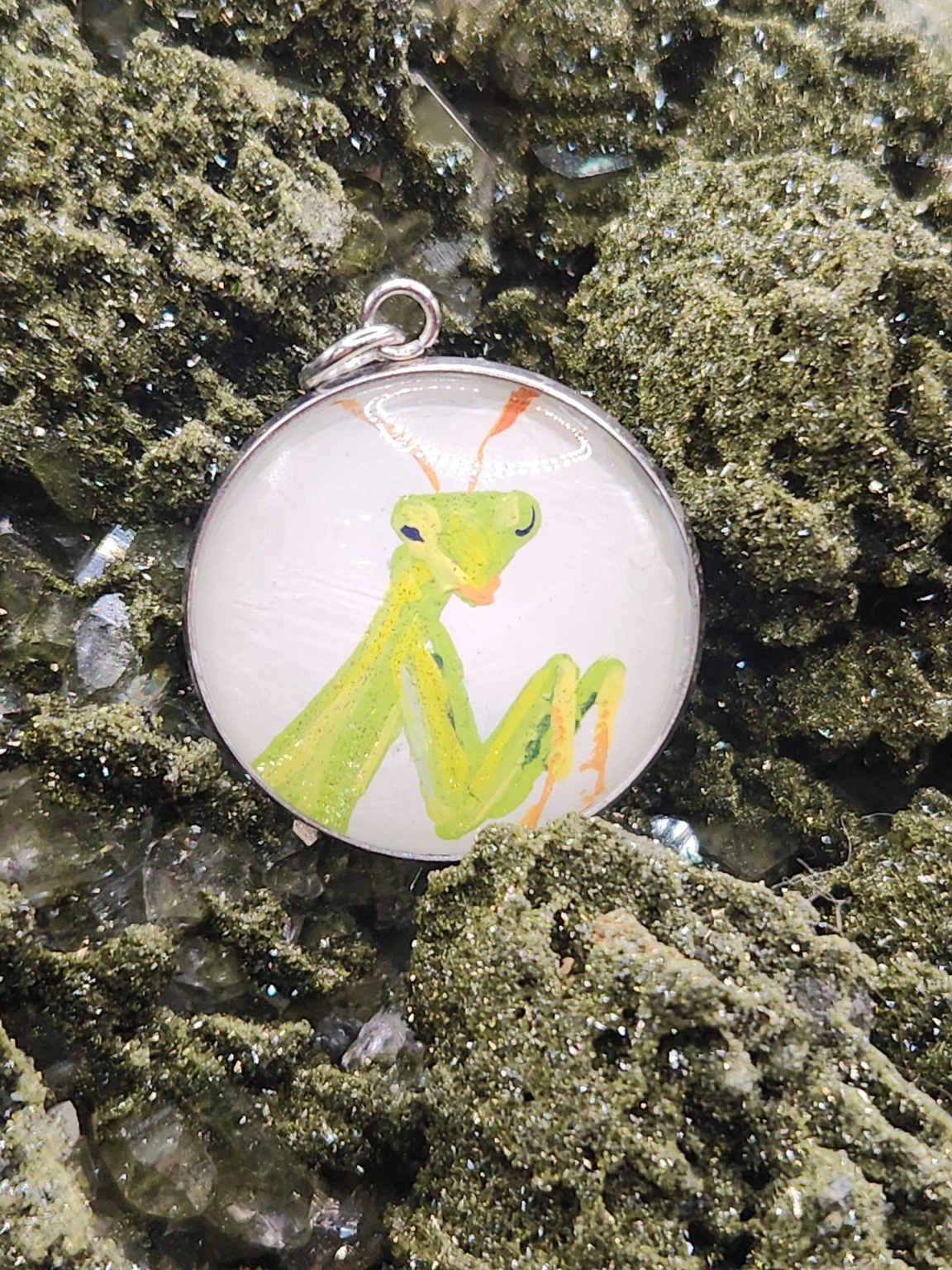 Green praying mantis art pendant resting on a green crystal display. Crystal display not included.
