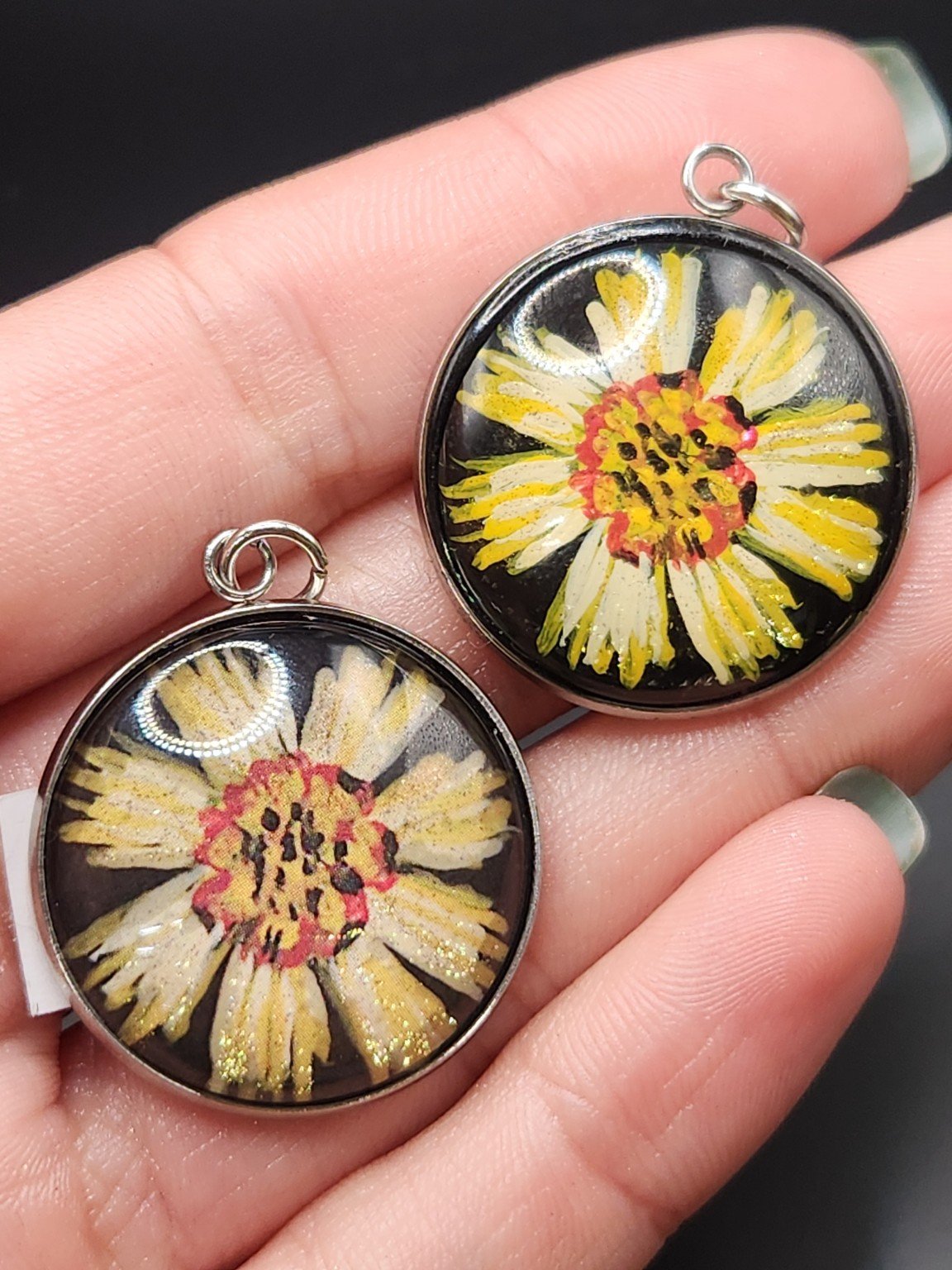 Two coreopsis flower pendants held in a hand: hand-painted original at the top right and printed reproduction at the bottom left.