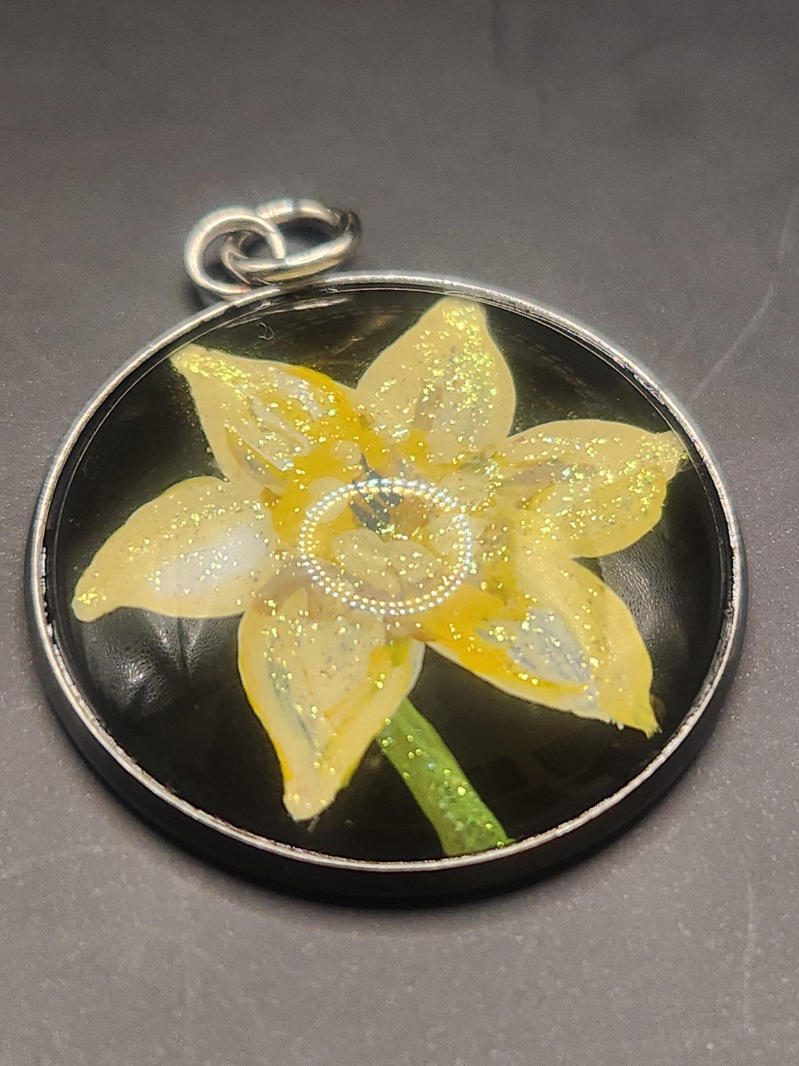 Yellow daffodil flower art pendant with shimmering glitter in a silver-tone bezel against a gray background.