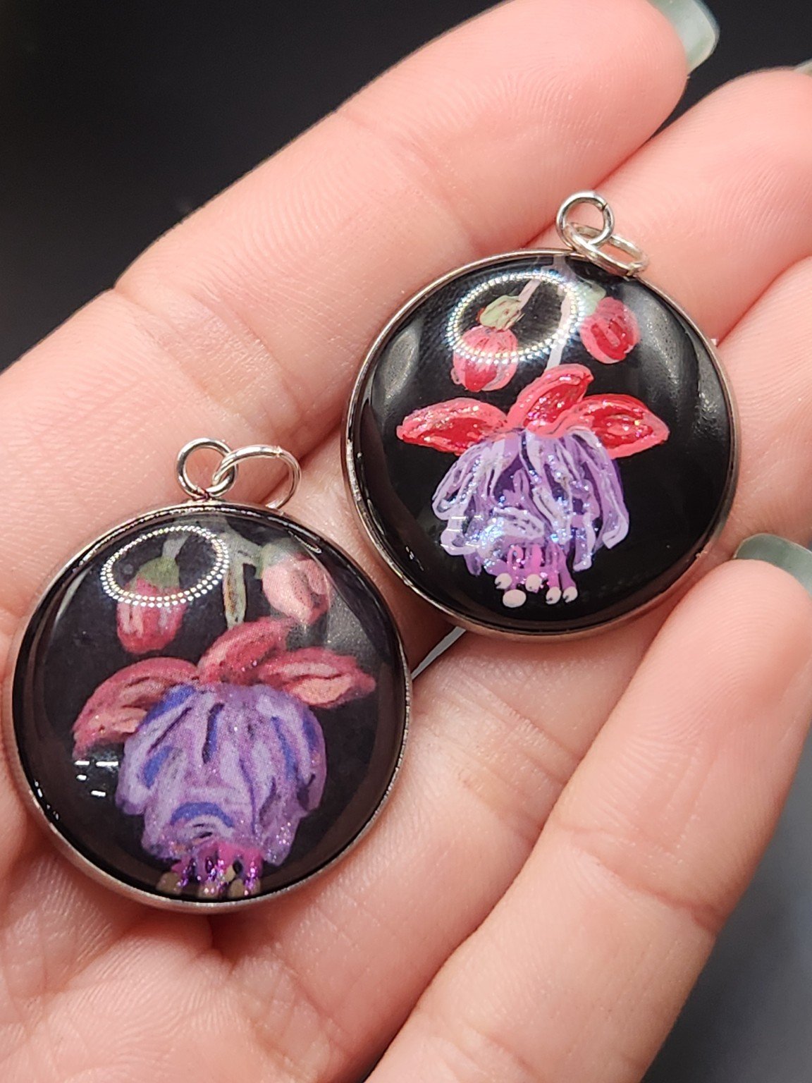 Two fuchsia flower pendants held in a hand: hand-painted original at the top right and printed reproduction at the bottom left.