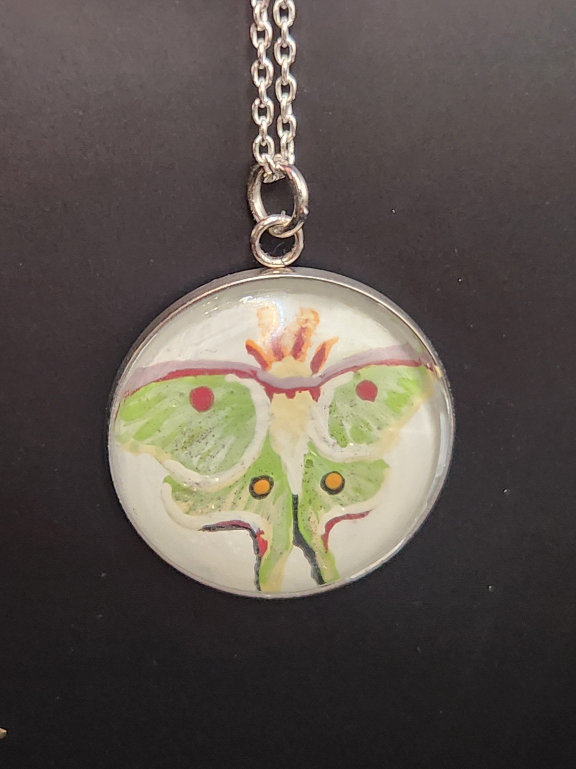Pale green luna moth art pendant shown on a silver-tone chain against a dark background. Chain not included.