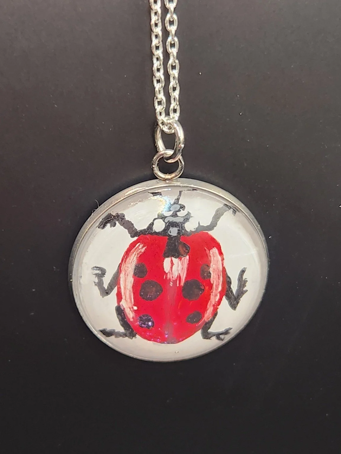 Red and black ladybug art pendant shown on a silver-tone chain against a dark background. Chain not included.