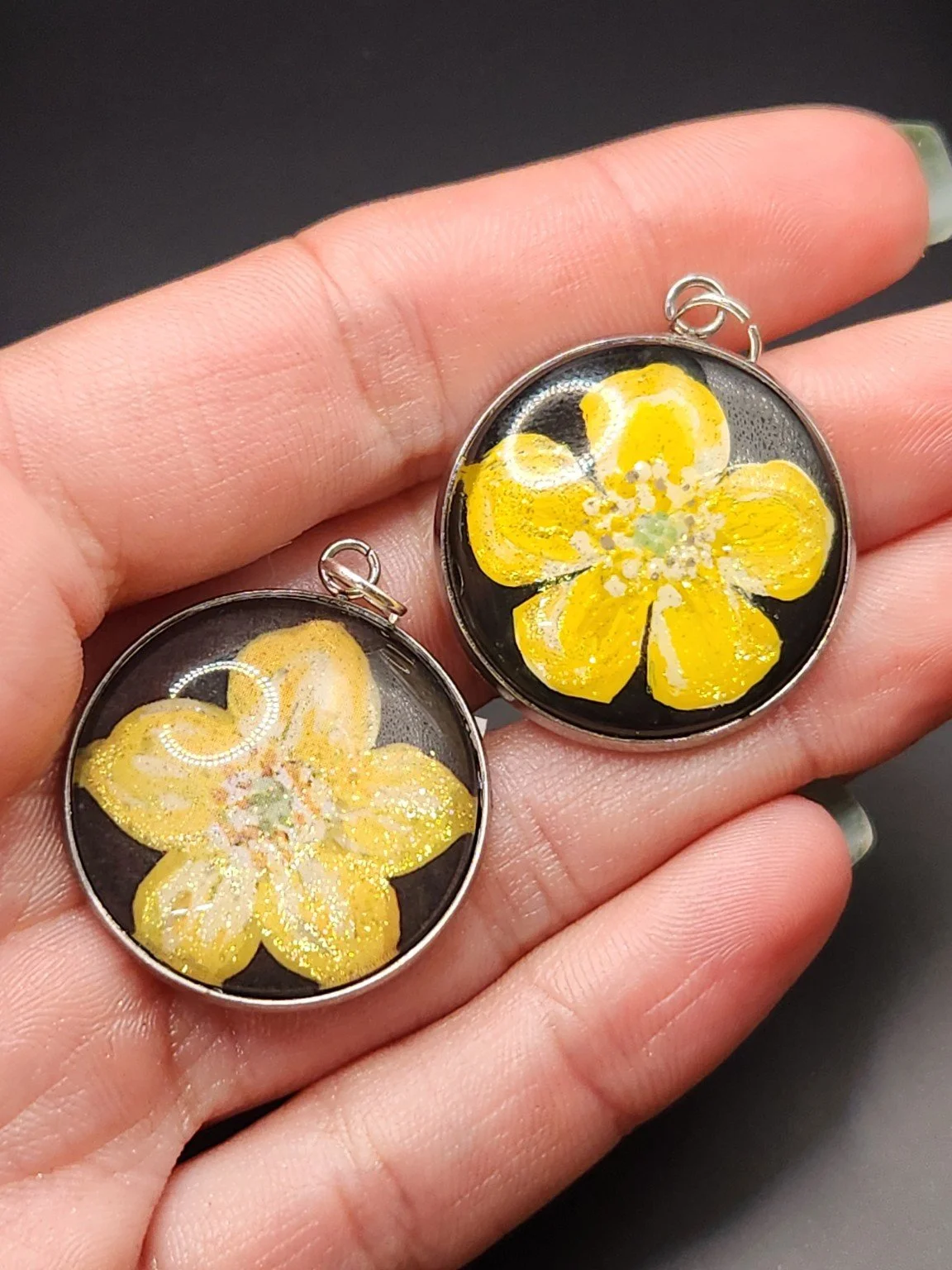 Two buttercup flower pendants held in a hand: hand-painted original at the top right and printed reproduction at the bottom left.