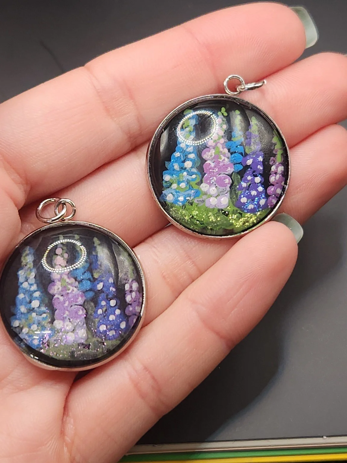Delphinium pendant comparison held in a hand: the hand-painted original is at the top right, and the printed reproduction is at the bottom left.