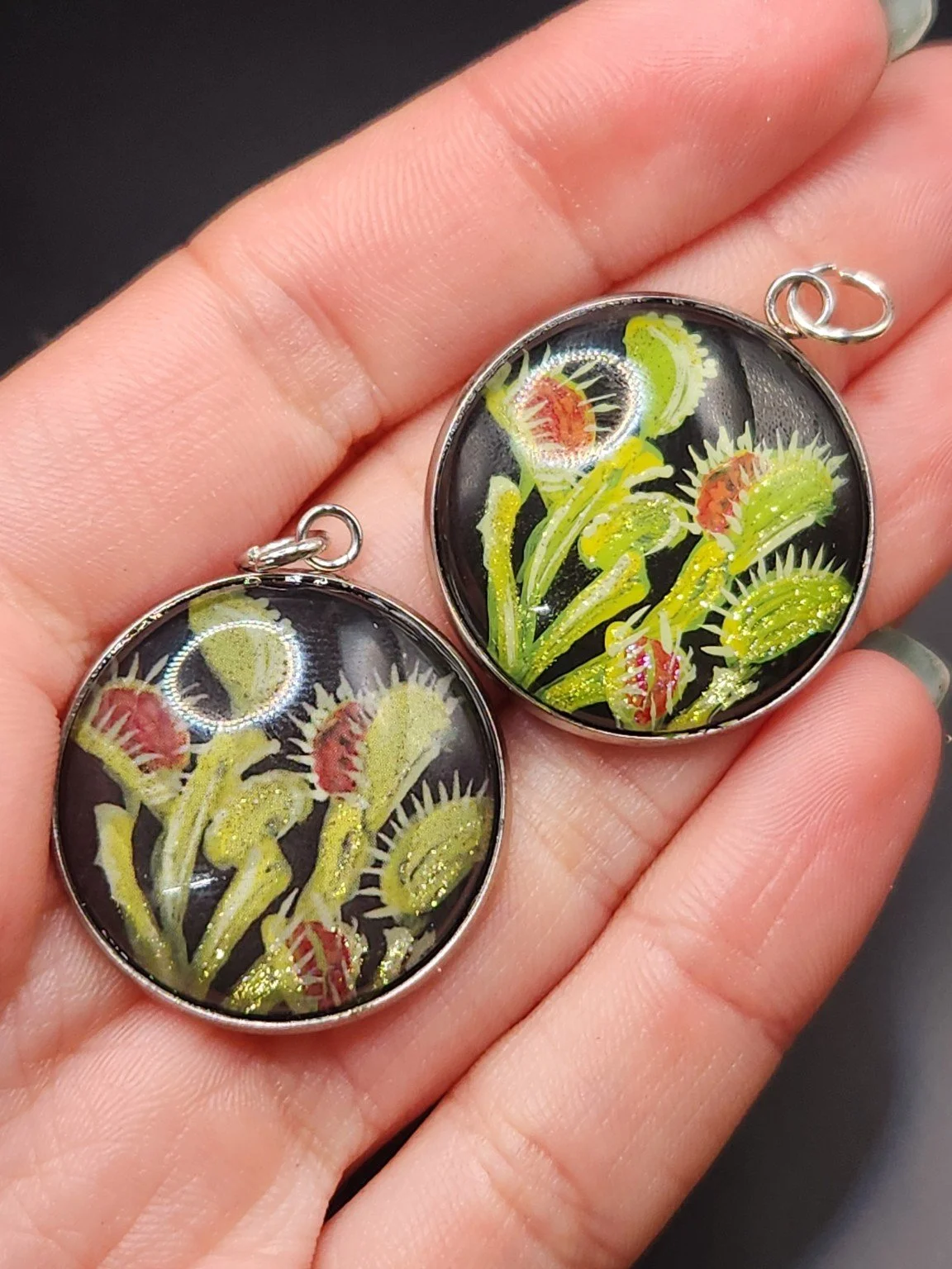 Two Venus flytrap art pendants held in a hand: hand-painted original at the top right and printed reproduction at the bottom left.