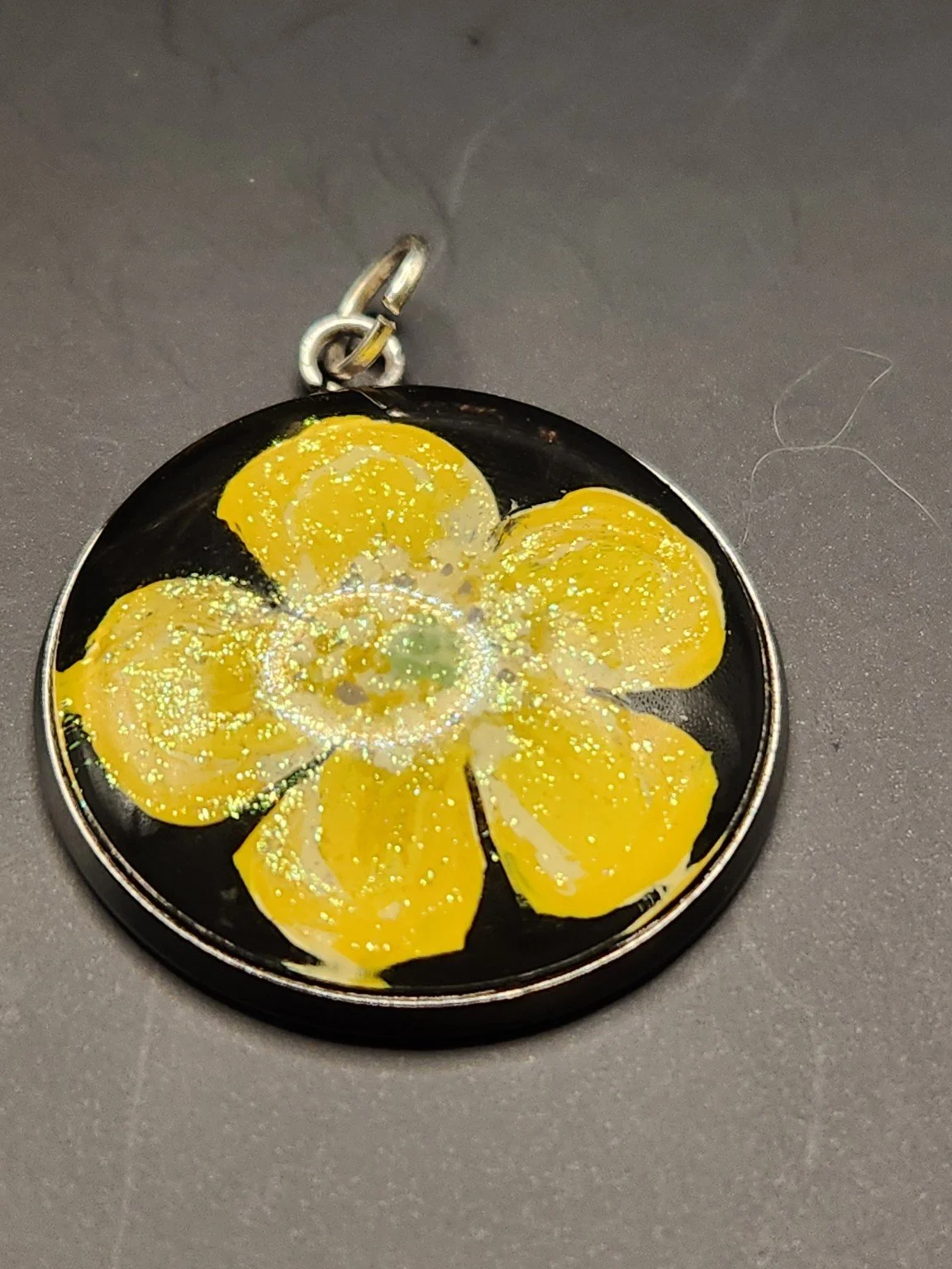 Golden yellow buttercup flower art pendant with shimmering glitter in a silver-tone bezel against a gray background.