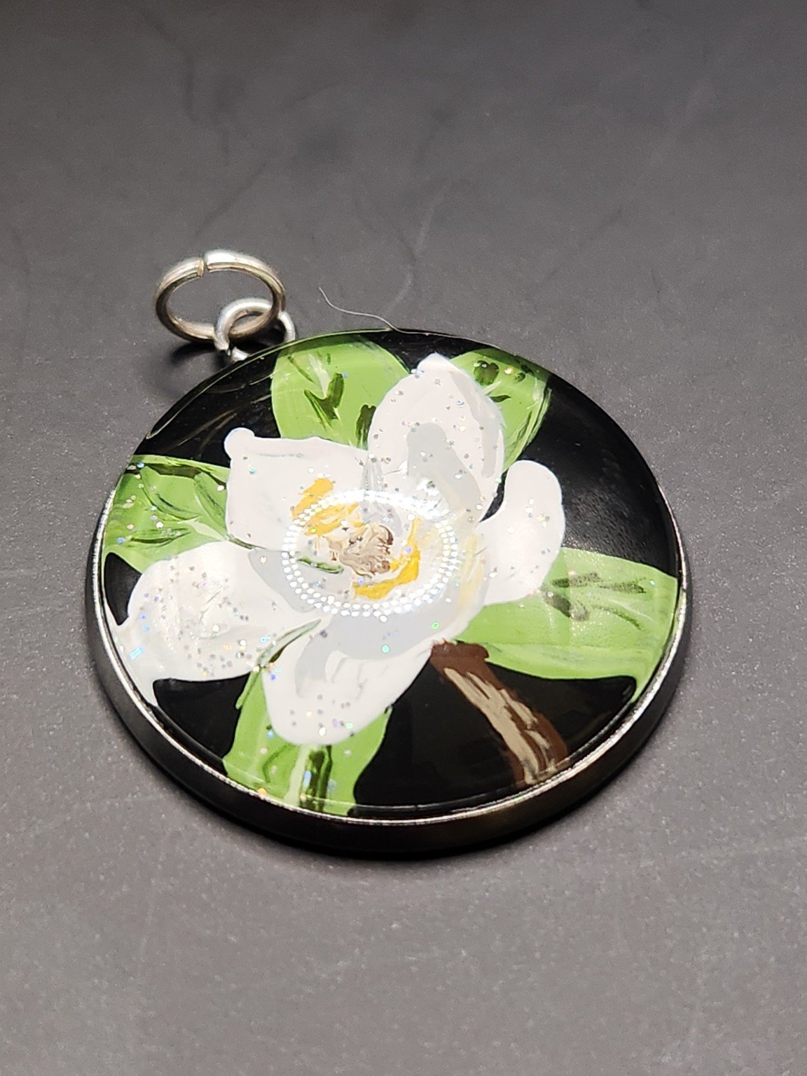 White magnolia flower art pendant with green leaves and glitter accents in a silver-tone bezel.