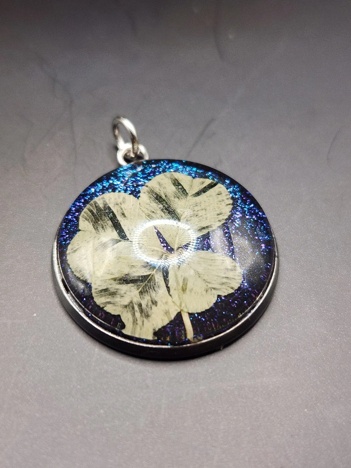 Pristine P002: real pressed four-leaf clover pendant with blue glitter background, close-up on gray.