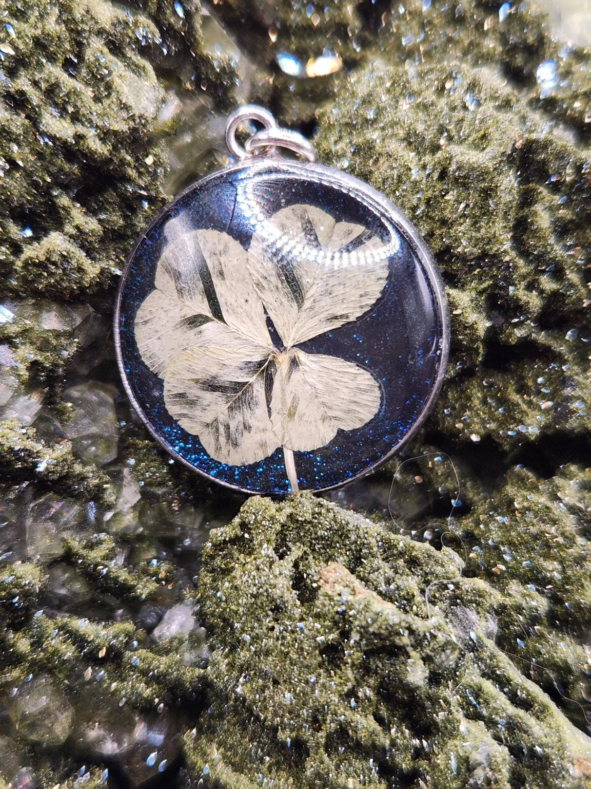 Pristine P002: real four-leaf clover pendant with blue glitter, shown on a crystal display. Display not included.