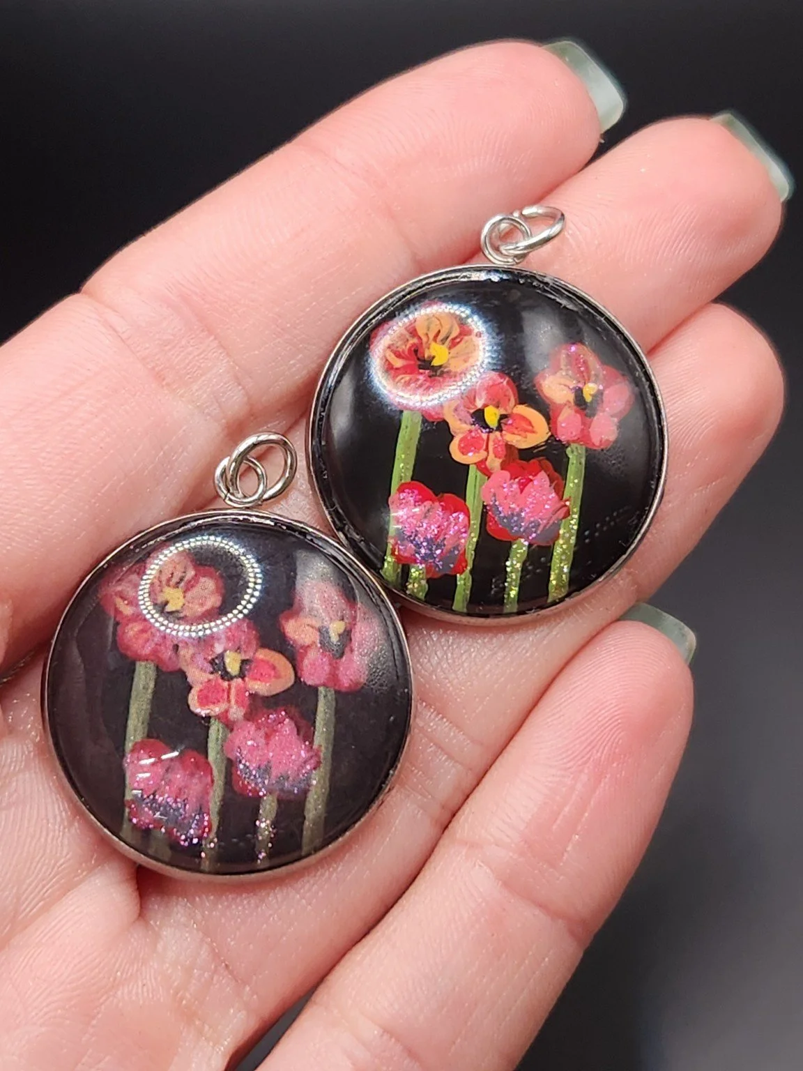 Two poppy flower pendants held in a hand: hand-painted original at the top right and printed reproduction at the bottom left.
