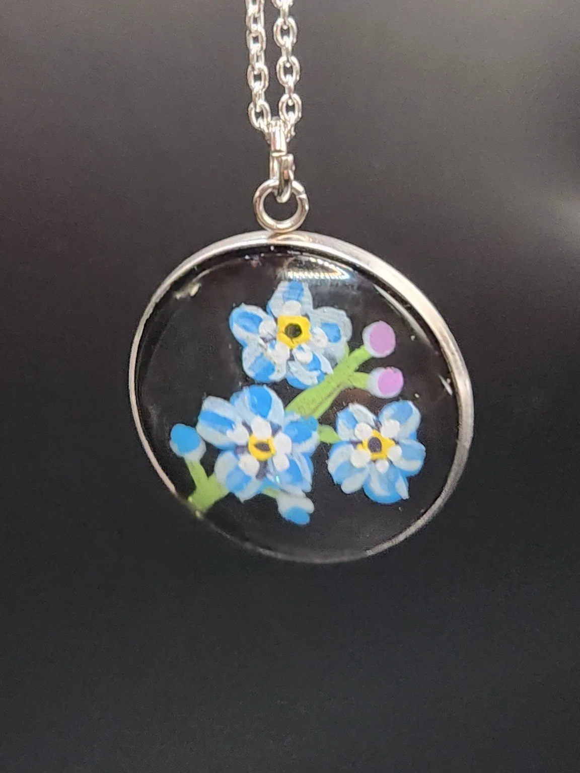Blue forget-me-not flower art pendant hanging from a silver-tone chain against a dark background. Chain not included.