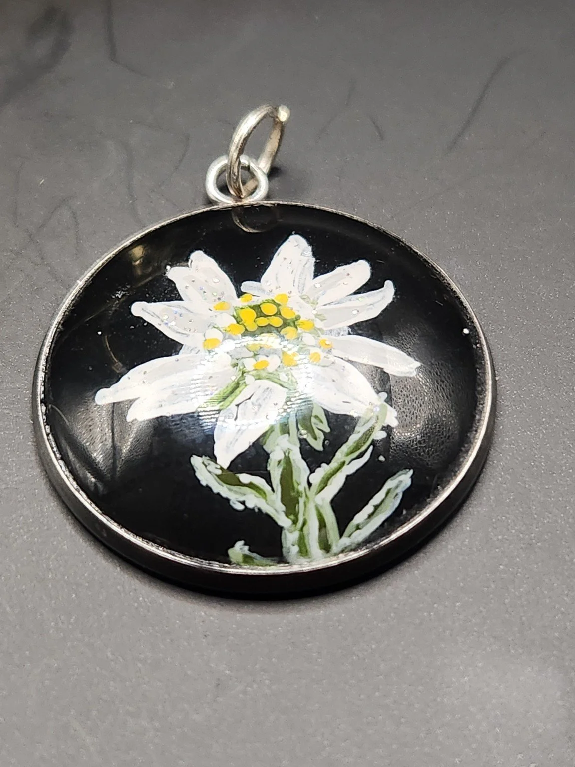 White edelweiss flower art pendant with yellow centers, green stems, and glitter accents on a gray background.