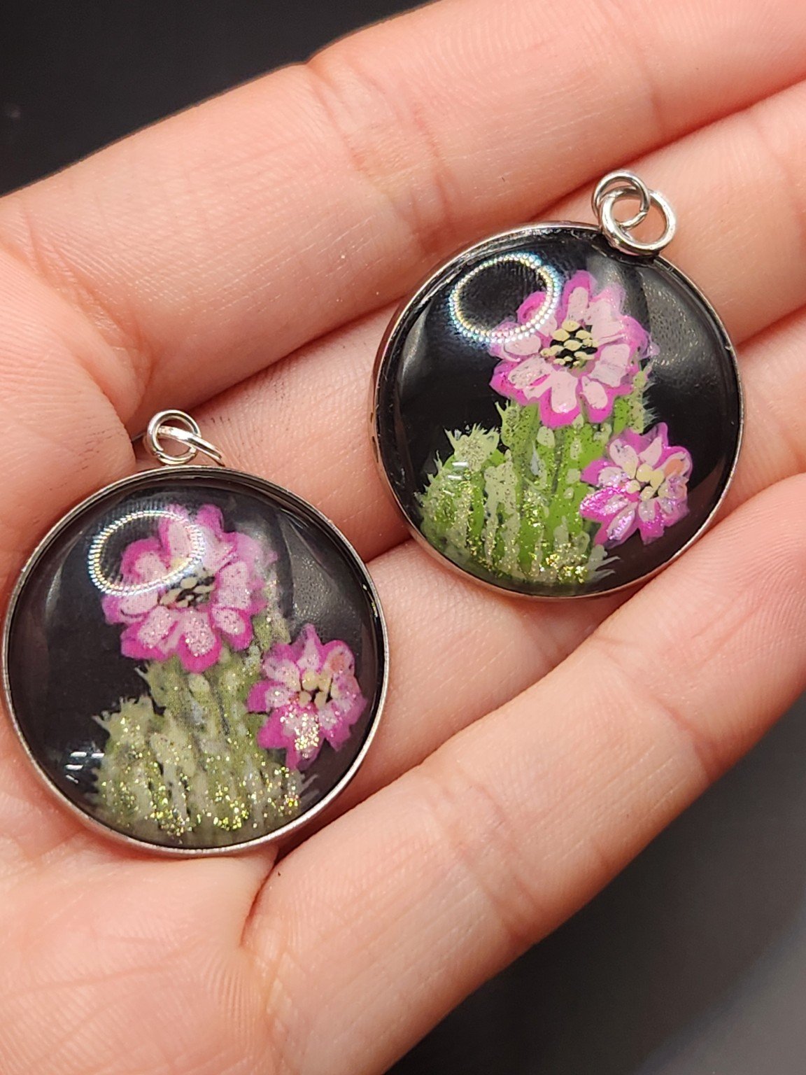 Two cactus flower pendants held in a hand: hand-painted original at the top right and printed reproduction at the bottom left.