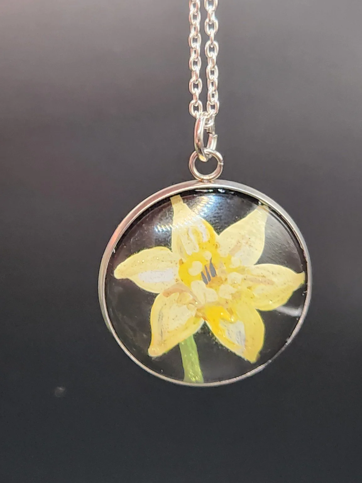 Yellow daffodil flower art pendant hanging from a silver-tone chain against a dark background. Chain not included.