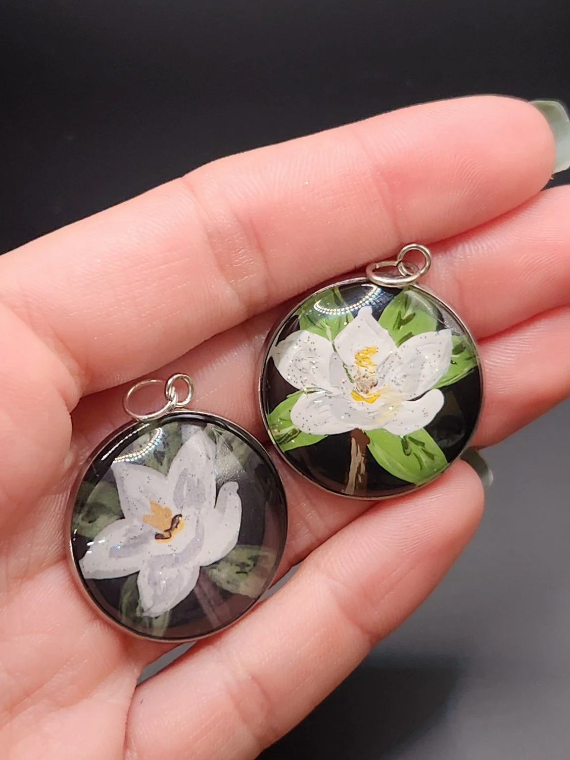 Two magnolia flower pendants in a hand: hand-painted original at the top right and printed reproduction at the bottom left.
