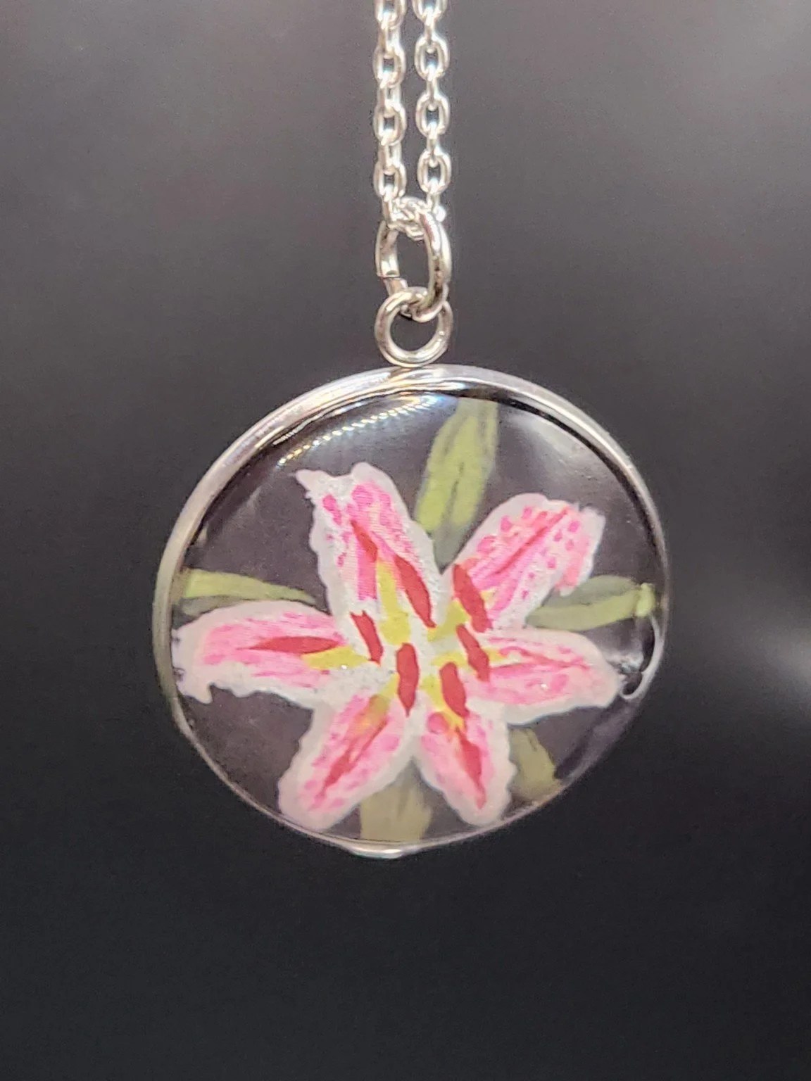 Pink-and-white Stargazer lily art pendant hanging from a silver-tone chain against a dark background. Chain not included.