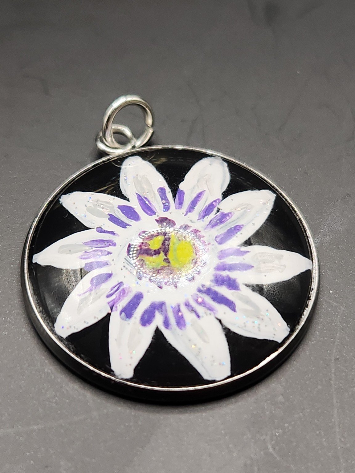 Purple and white passion flower art pendant with a yellow-green center and glitter accents in a silver-tone bezel against a gray background.
