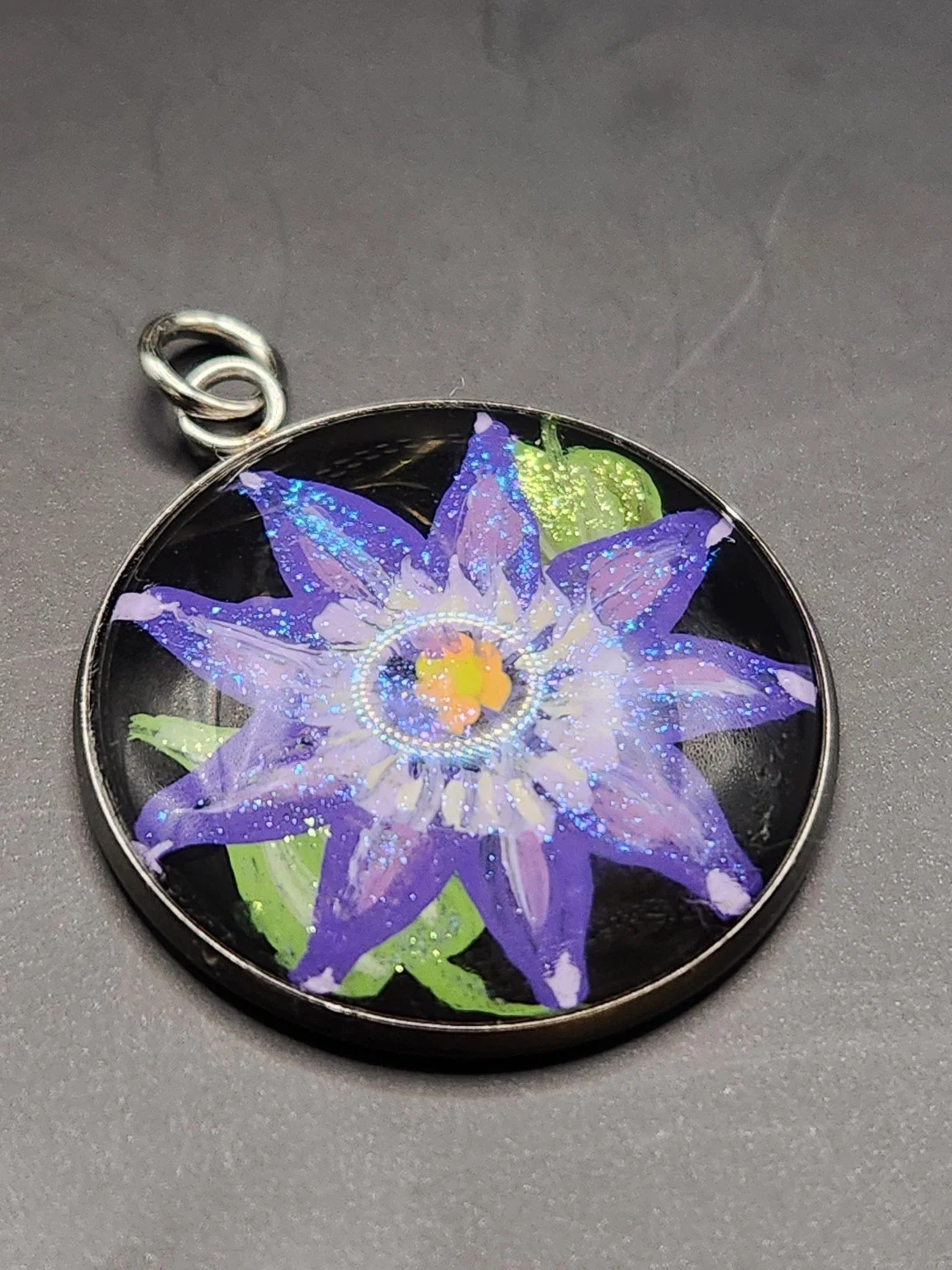 Purple clematis flower art pendant with glitter accents in a silver-tone bezel on a gray background.