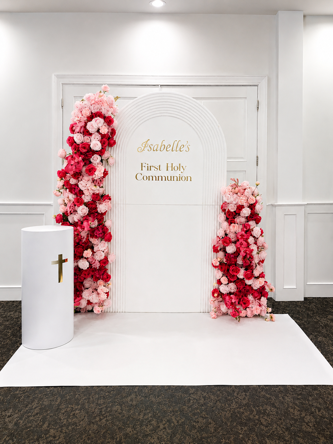 A white backdrop with gold lettering reads "Isabelle's First Holy Communion," decorated with pink and red floral arrangements on both sides and a white pedestal with a gold cross in front. Chicago events decor photo booth rental