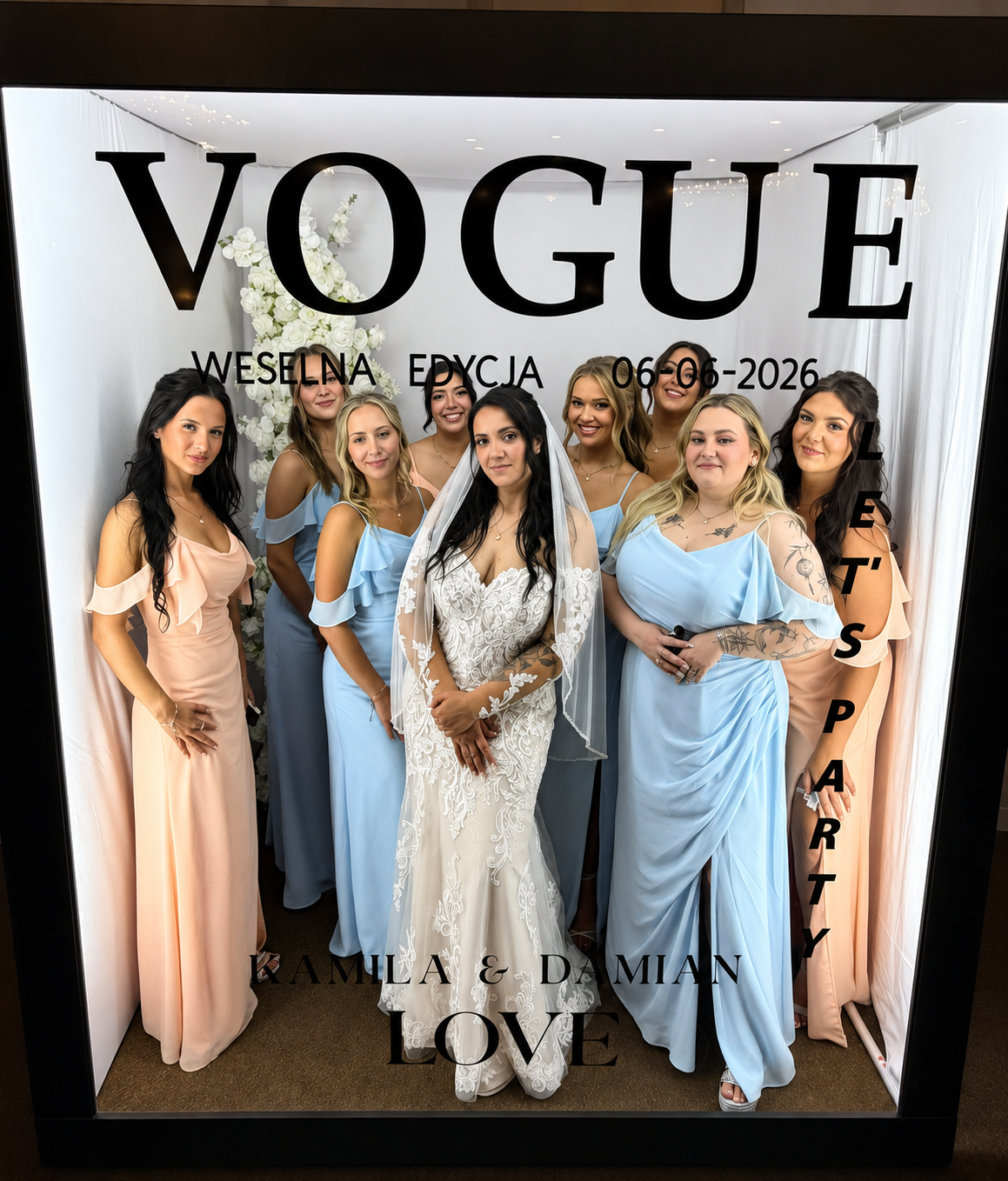 A group of women at a wedding, with the bride in a lace wedding gown and veil, surrounded by her bridesmaids in pastel-colored dresses. They are posing in front of a photo booth with a floral backdrop and the words "VOGUE" and event details written o